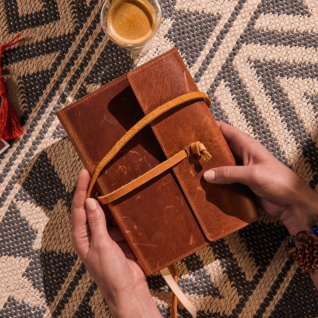 Brown leather journal with a strap on a patterned surface with a cup and photos.