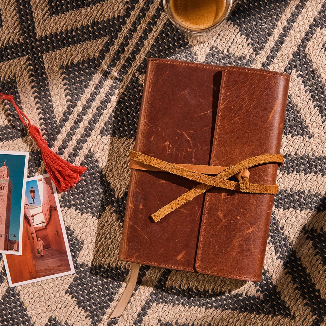 Brown leather journal with a strap on a patterned surface with a cup and photos.