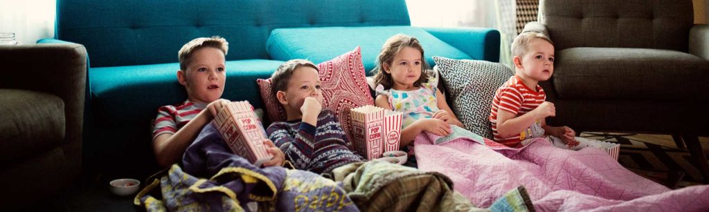 Kids sitting on a couch watching a movie with blankets and popcornn